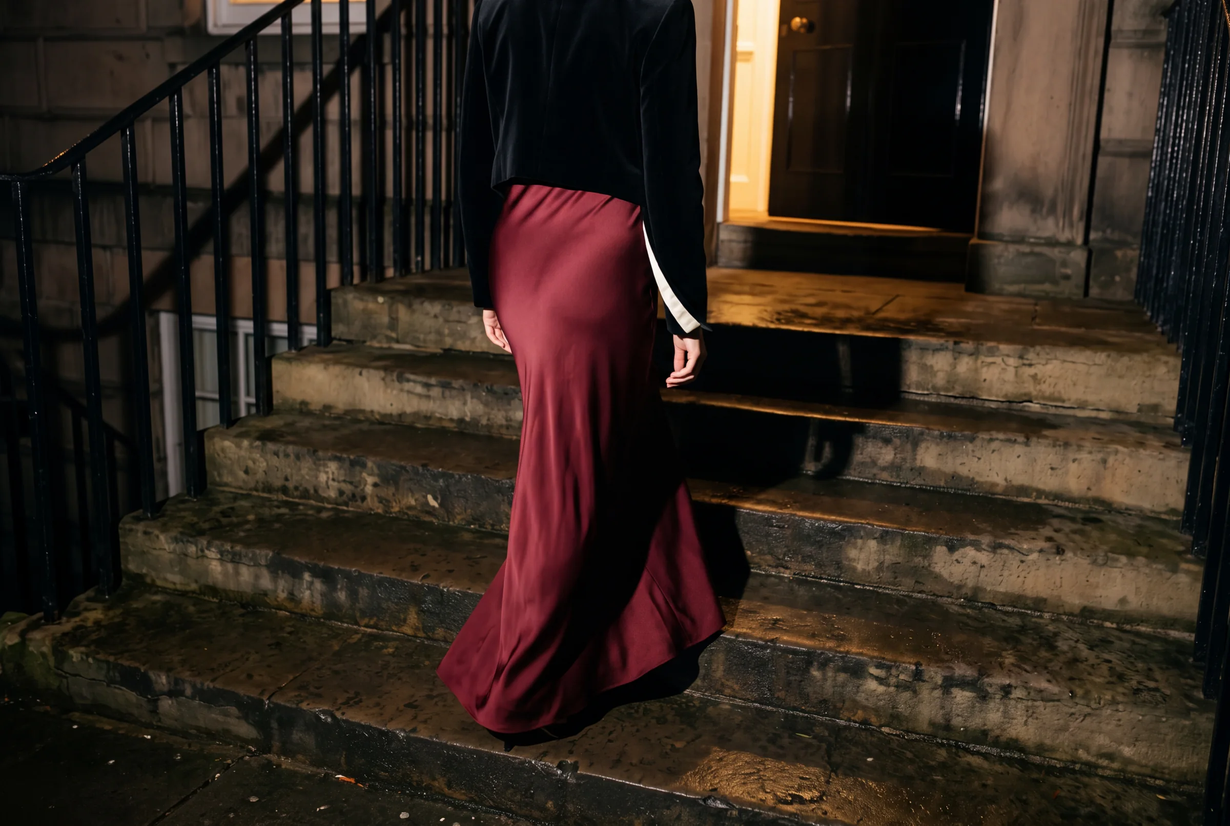 A close fashion-editorial crop of an oxblood silk dress moving up rain-darkened limestone steps at night