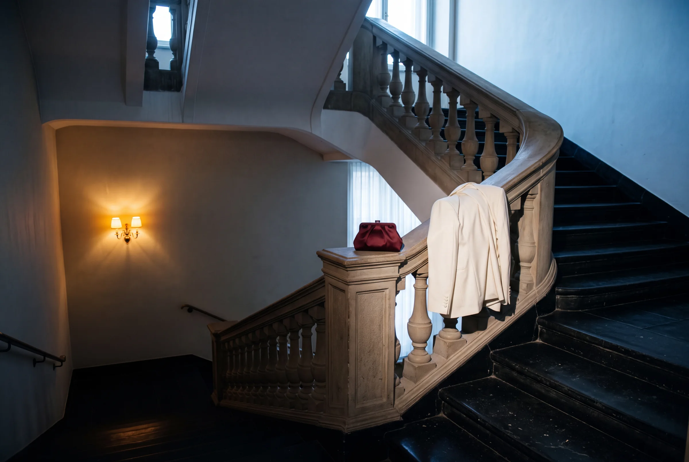 An empty hotel stairwell moving from warm night light into cool dawn with an ivory coat and oxblood evening bag